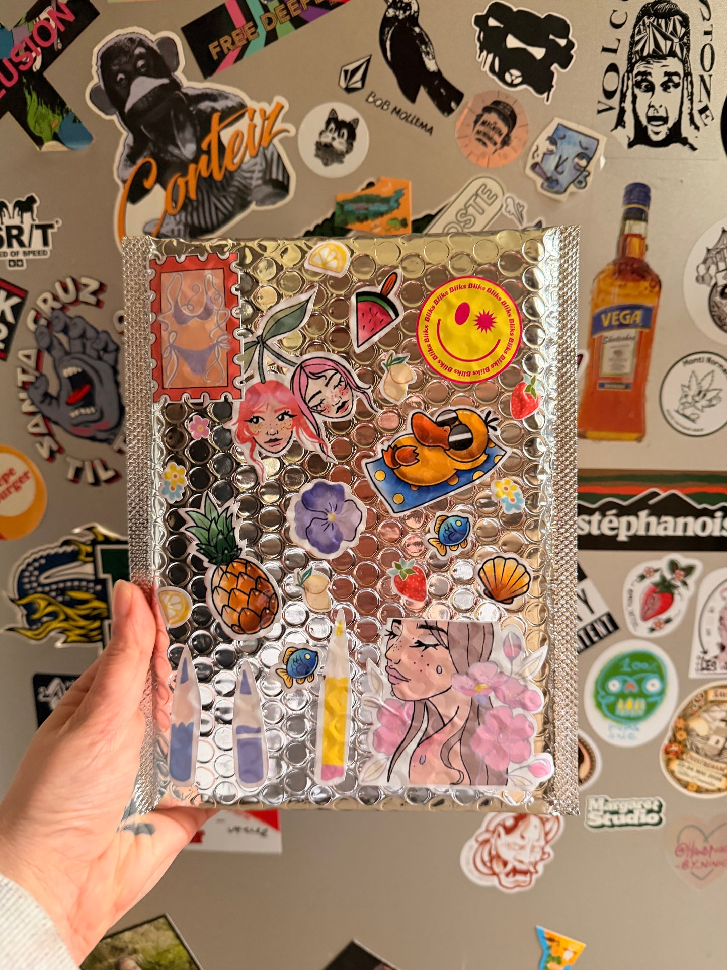 Pochette argentée remplie de stickers illustrés colorés et packaging personnalisé Bliks devant un mur de stickers.