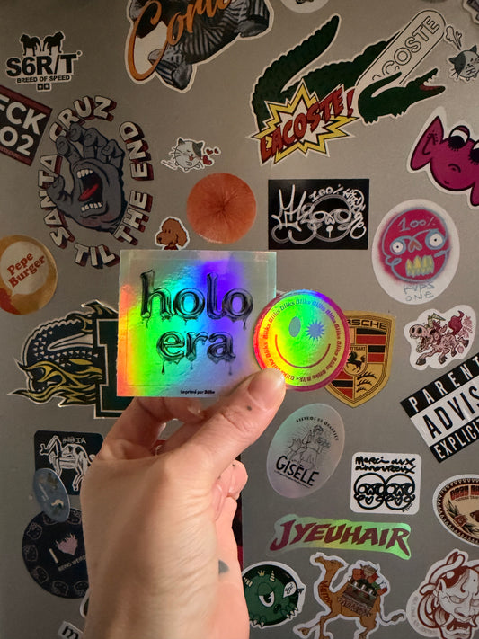Deux stickers holographiques personnalisés avec effet miroir arc-en-ciel tenus à la main devant un mur de stickers.